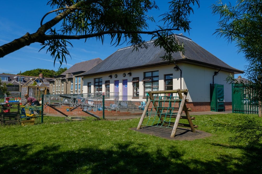 A single-level schoolbuilding surrounded by a green fence, also containing a playground.