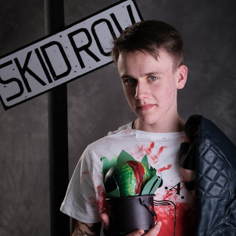 From the show Little Shop of Horrors, an actor in a white bloodstained t-shirt with a leather jacket over his shoulder. He is holding a small prop plant - little Audrey 2.