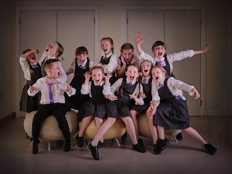 11 rebellious children in school uniform are posed cheekily in a group, as characters from the show Matilda.