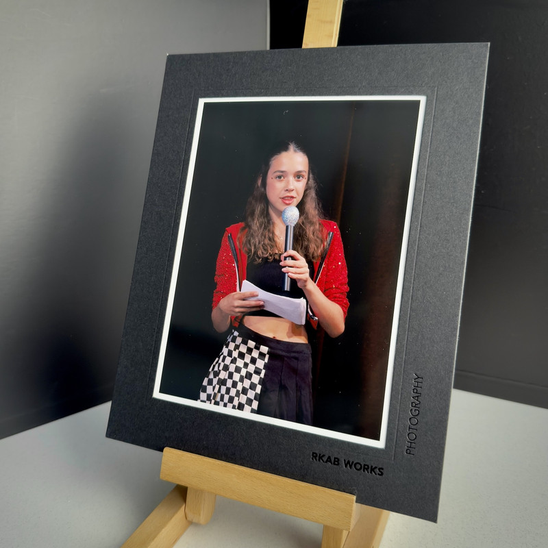 An actress in a checkered skirt and sparkly red jacket reads from a card into a sparkly microphone. The photo is printed in and presented in a black frame, presented on a small desktop easel.