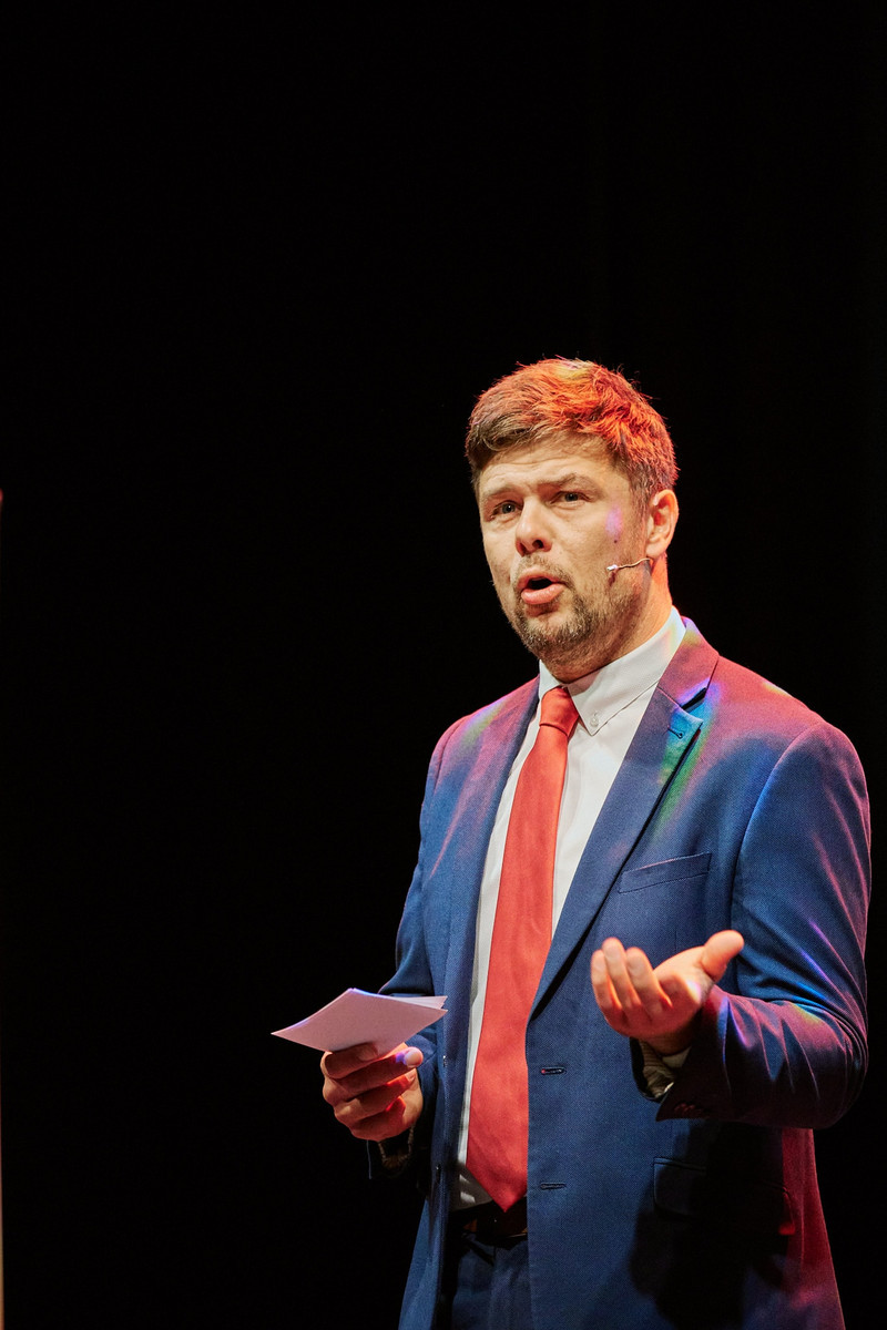 A man in a blue suit jacket with a red tie presents on a stage.