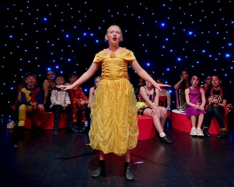 A young actress in a yellow dress sings with her hands out to her sides. Behind her there are other costumed children and a starry background.