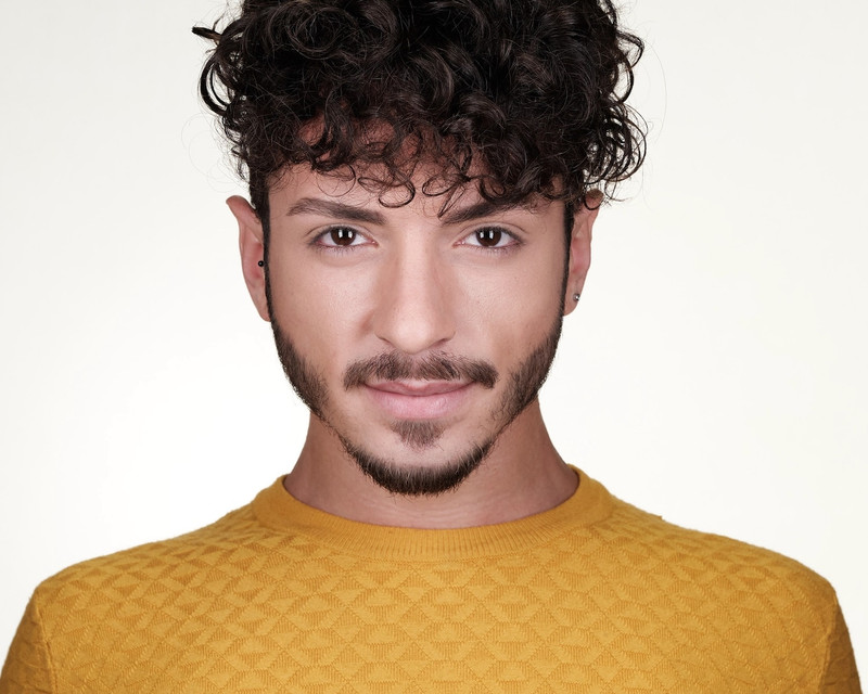 In a yellow jumper, a curly-haired man looks sharply to the camera.