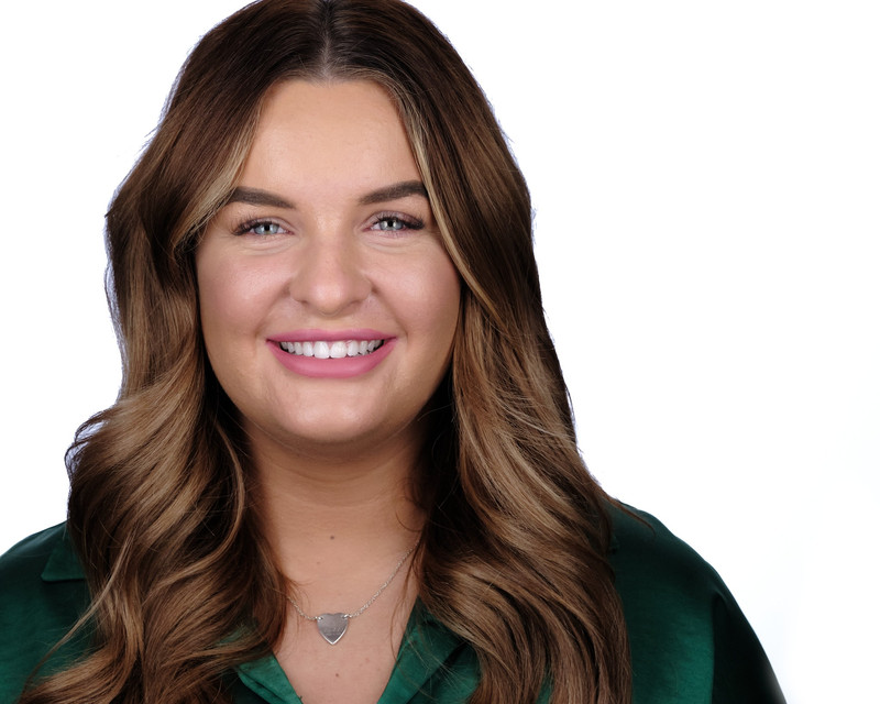 A woman with waves in her hair smiles to the camera. She is wearing a silver heart-shaped necklace, just above a green collared top.