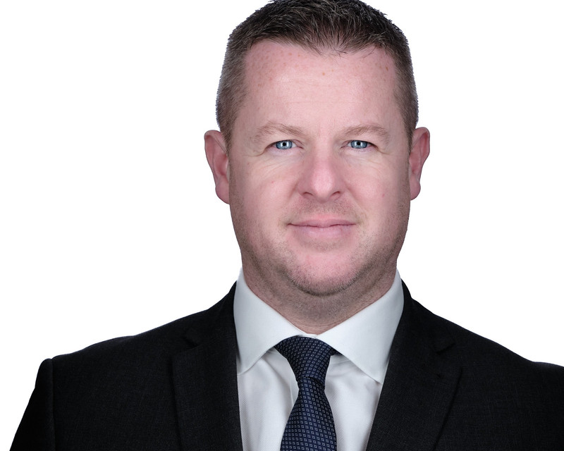 A short-haired main wears a suit with a blue patterned tie, he looks confidently to the camera with blue eyes.