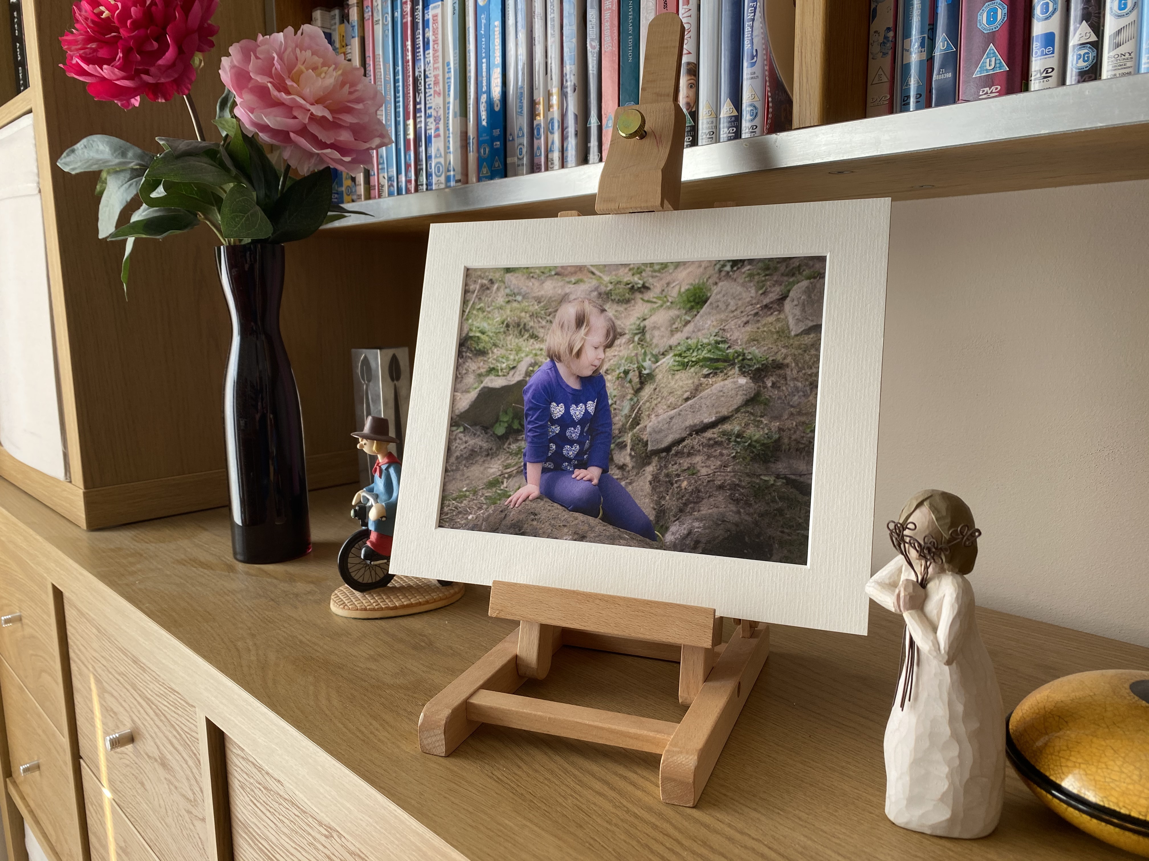 A child wearing blue and looking to the side, sitting on a rock. The image is in a light-coloured, slim mounting frame and in an easel, surrounded by ornaments.